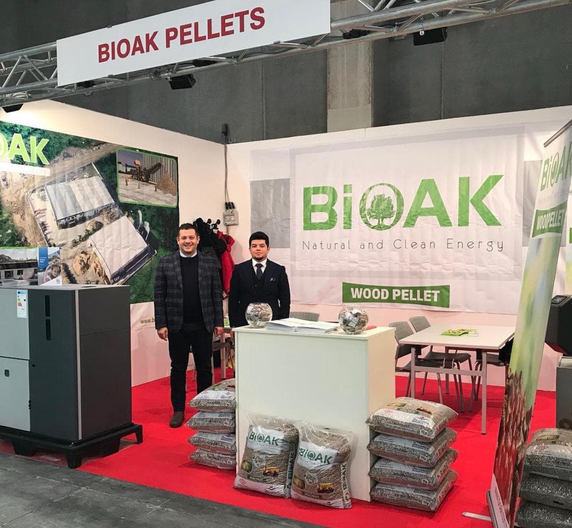 İki adam, istiflenen çantalar ve ekipmanlarla bir BiOAK ahşap pelet fuarı standında.