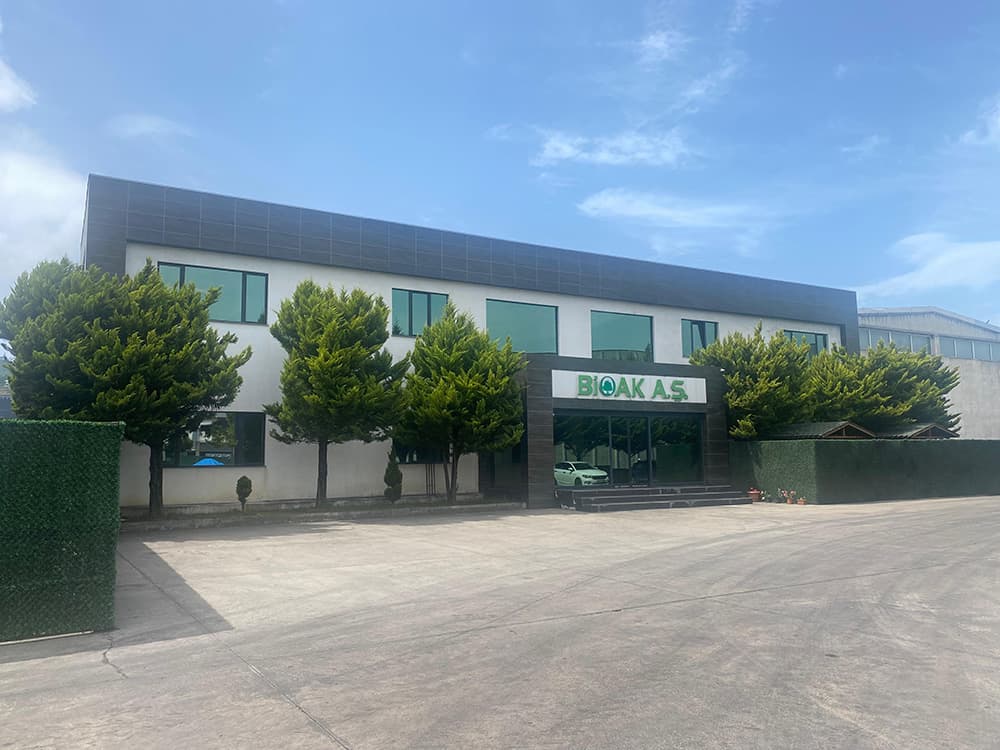 Modern two-story office building with BIOAK A.S. signage, green windows, and landscaped evergreen trees.